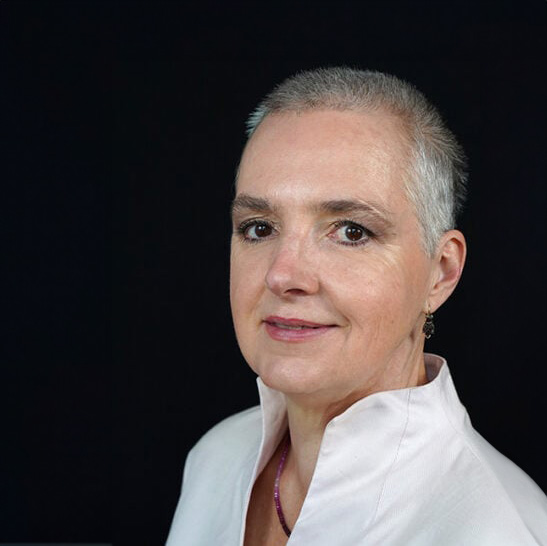Eine Frau mit kurzen grauen Haaren, einem weißen Oberteil mit Kragen und kleinen Ohrringen posiert vor einem schwarzen Hintergrund. Sie blickt leicht zur Seite und lächelt sanft in die Kamera.