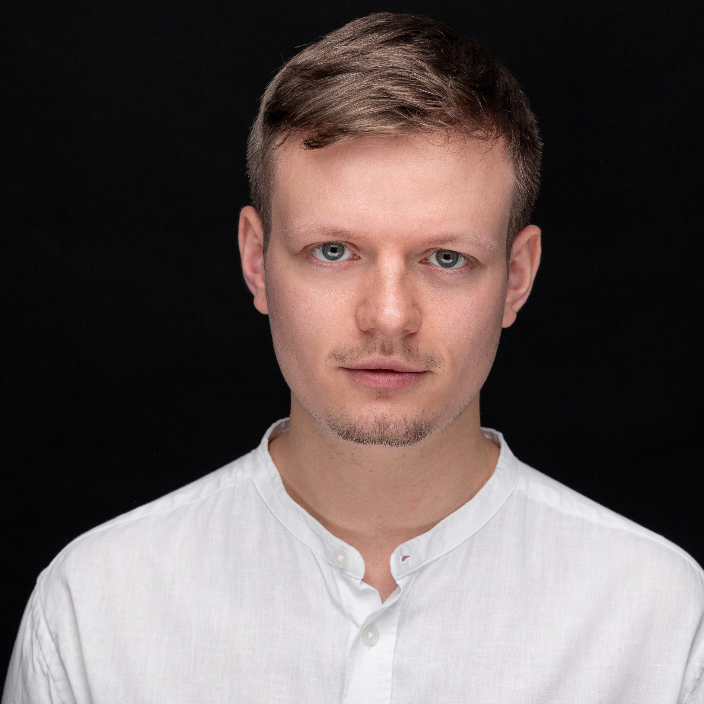 Ein junger Mann mit kurzen hellbraunen Haaren und blauen Augen, der ein weißes Hemd mit Mandarinkragen trägt, blickt vor einem schlichten schwarzen Hintergrund direkt in die Kamera - genau richtig für eine Veranstaltung wie Musik in der St. Marienkirche.