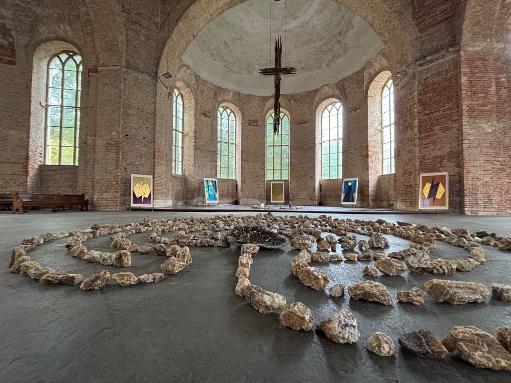 Große kreisförmige Steinmuster auf dem Boden eines geräumigen, gemauerten Raums mit hohen Bogenfenstern; Kunstwerke sind auf Staffeleien an den Wänden ausgestellt.