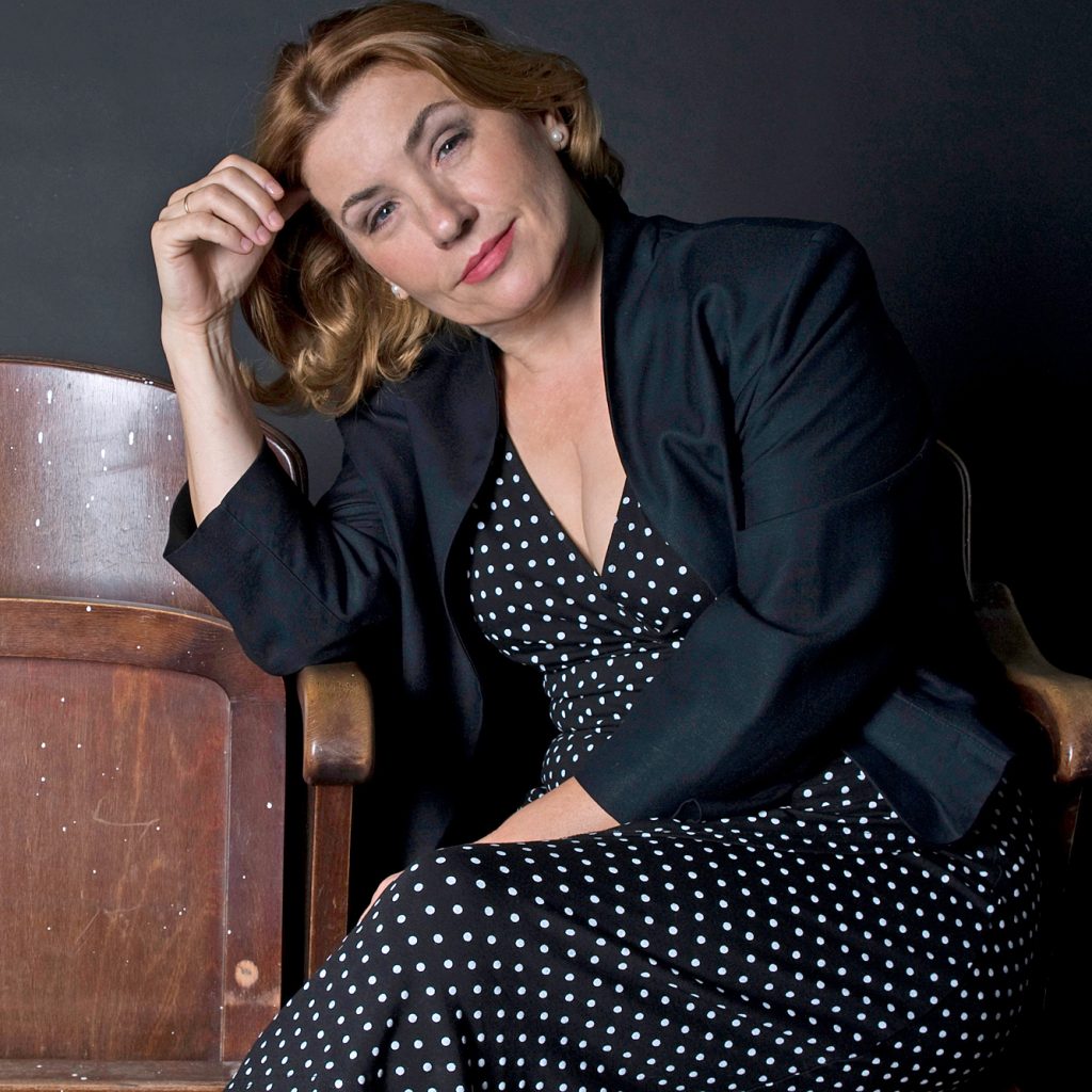 Eine Frau mit hellbraunem Haar, die ein schwarzes gepunktetes Kleid und einen schwarzen Blazer trägt, sitzt entspannt auf einem Holzstuhl, stützt ihren Kopf auf die Hand und schaut in die Kamera vor einem dunklen Hintergrund.