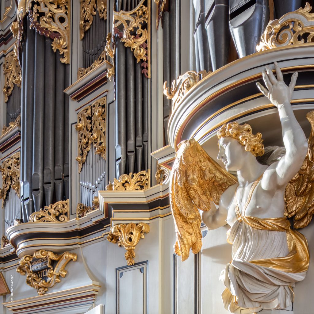 Nahaufnahme einer kunstvollen Pfeifenorgel mit goldenen Details und einer skulptierten Engelsfigur mit goldenen Flügeln vor einem komplizierten Hintergrund aus Pfeifen und kunstvollen Dekorationen.