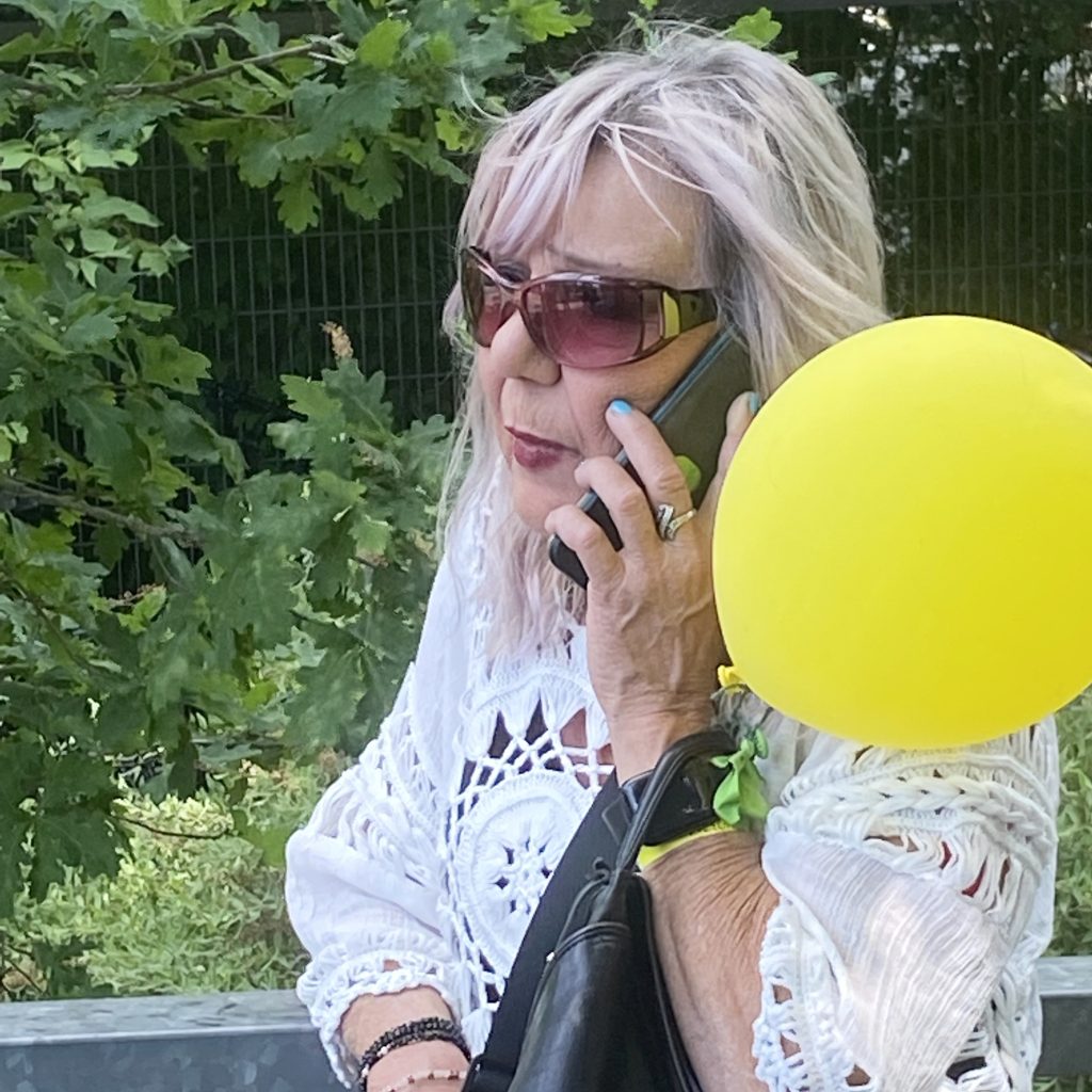 Eine Frau mit hellem Haar und Sonnenbrille spricht im Freien mit einem Handy. Sie trägt ein weißes Häkeltop, hat eine schwarze Tasche in der Hand und trägt einen gelben Luftballon am Handgelenk. Im Hintergrund ist grünes Laub zu sehen.