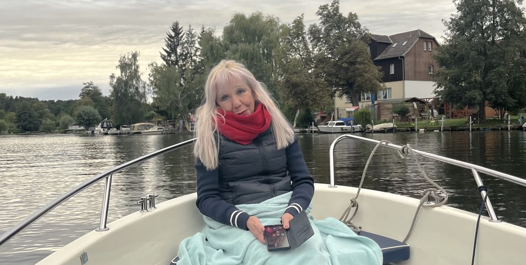 Eine Frau mit langen blonden Haaren, einem roten Schal und einer dunklen Weste sitzt auf einem Boot und hält ein Telefon in der Hand. Sie ist in eine hellblaue Decke gehüllt. Im Hintergrund sind ein ruhiger Fluss und von Bäumen umgebene Häuser zu sehen.