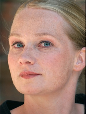 Eine Frau mit heller Haut, blauen Augen und blondem, zurückgegeltem Haar blickt leicht zur Seite. Sie hat Sommersprossen und einen neutralen Gesichtsausdruck. Sie trägt ein schwarzes Oberteil mit unscharfem Hintergrund.