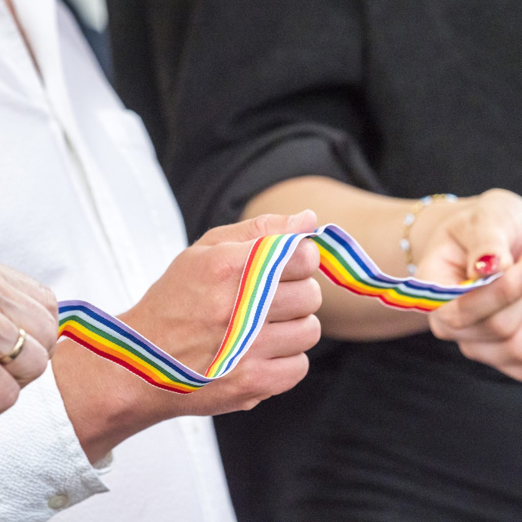 Drei Menschen halten gemeinsam ein regenbogenfarbenes Band in den Händen, das für Einheit und Vielfalt steht. Zu sehen sind nur ihre Hände und ein Teil ihres Oberkörpers.