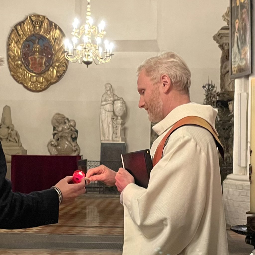 Ein Mann in einem weißen Gewand nimmt in einem prunkvollen, von Kerzen beleuchteten Raum, der mit Statuen und religiösen Kunstwerken geschmückt ist, einen roten Apfel von einer anderen Person entgegen.